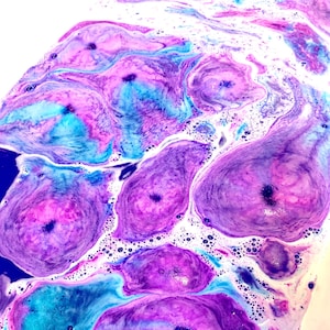 May include: Close-up of a bath bomb dissolving in water, creating a swirling pattern of colours. The colours include shades of purple, blue, and pink, with white foam and bubbles. The bath bomb is creating a marbled effect.