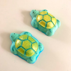 May include: Two green bath bombs shaped like turtles with yellow shells. The bath bombs have a sparkly finish.