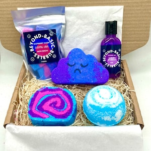 May include: A gift box containing bath products. It features a bag of bath rocks, a purple shower gel bottle, and three bath bombs in different shapes and colours. The bath bombs are blue, pink, and purple. The box is lined with straw and white paper.