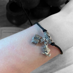 May include: A black cord bracelet with a silver charm featuring a witch on a broomstick and a book. The charm is attached to a silver bead with floral designs.
