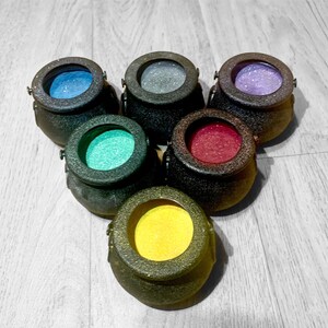 May include: Six small black cauldron-shaped containers with different colored glitter inside. The cauldrons are arranged in a circle.