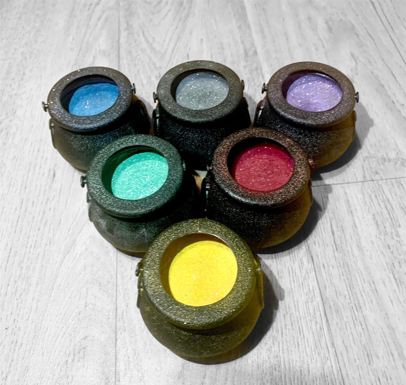 May include: Six small black cauldron-shaped containers with different colored glitter inside. The cauldrons are arranged in a circle.