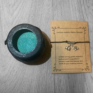 May include: A black cauldron charm bracelet with silver charms of an open book and a wizard's hat. The bracelet is on a brown card with the text "Potions master charm bracelet" and "It takes a truly great wizard to produce such magnificent potions! Wear this bracelet as a mark of your magical talents."