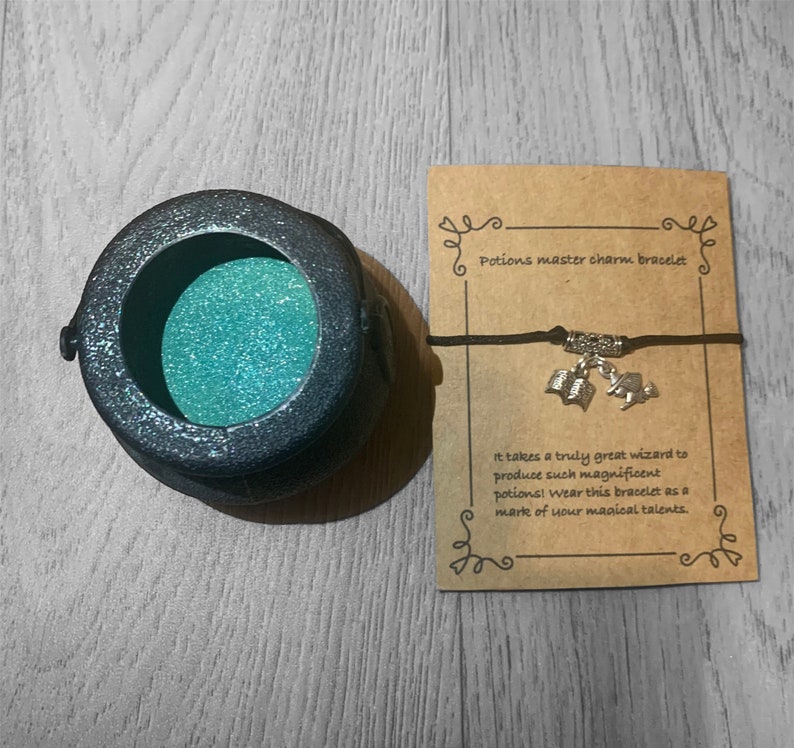 May include: A black cauldron charm bracelet with silver charms of an open book and a wizard's hat. The bracelet is on a brown card with the text "Potions master charm bracelet" and "It takes a truly great wizard to produce such magnificent potions! Wear this bracelet as a mark of your magical talents."