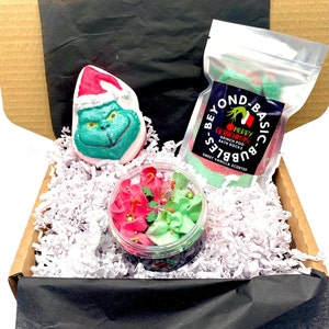 May include: A festive gift box featuring a Grinch-shaped bath bomb, a container of red and green bath frosting, and a bag of green and red bath rocks. The bath bomb is green with a red Santa hat. The bag of bath rocks has a label that reads "Merry Grinchmas".