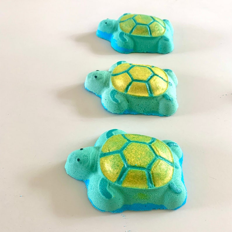 May include: Three turquoise bath bombs shaped like turtles with gold shells. The bath bombs have a blue rim around the edge.