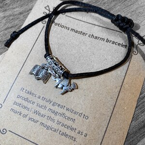 May include: Black string bracelet with silver charms, including a book, a bird, and a scroll. The bracelet is on a brown paper card with the text "Potions master charm bracelet" and "It takes a truly great wizard to produce such magnificent potions! Wear this bracelet as a mark of your magical talents."
