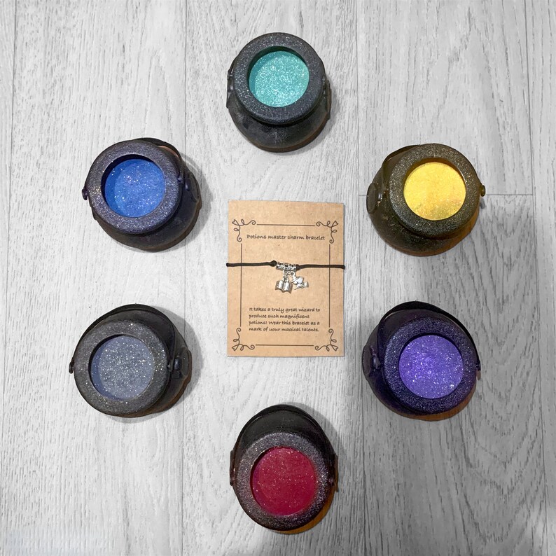 May include: Six small black cauldron-shaped containers with different colored glitter inside. The cauldrons are arranged in a circle around a card with the text "Potion master charm bracelet" and a charm bracelet with small charms.