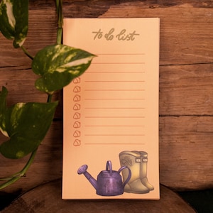 Puede incluir: Un bloc de notas de color melocotón para la lista de tareas con el título escrito a mano "to do list". El bloc de notas tiene 15 líneas para escribir tareas. El bloc de notas tiene una ilustración decorativa de una regadera y dos botas.