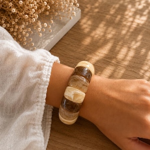 May include: A chunky bracelet with alternating sections of light and dark brown, worn on a wrist. The bracelet is made of rounded rectangular segments. The background includes a wooden surface and a white shirt.
