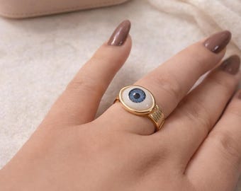 May include: Gold-toned ring featuring an eye design with a blue iris and white background. The ring has a textured band with small embellishments. Jewelry box in the background.