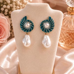 May include: A pair of earrings featuring a teal, spiral-shaped shell design with gold-toned accents and a central white pearl. Each earring includes a dangling, irregular-shaped white pearl drop suspended from the base of the shell component.