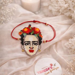 Könnte beinhalten: Ein Perlenarmband mit einem Porträt von Frida Kahlo. Das Porträt ist in einem pixeligen Stil mit roten, schwarzen und beigen Farbtönen gehalten. Das Armband hat eine rote Kordel und goldfarbene Akzente. Eine Kerze und Blumen sind im Hintergrund.
