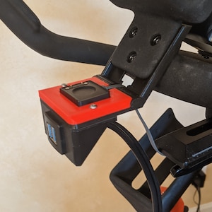 May include: A black and red 3D printed mount with two USB ports and a small black circular opening. The mount is attached to a black exercise bike.