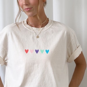 Op de afbeelding: Een wit T-shirt met een rij van vijf gekleurde harten die op de voorkant zijn gedrukt. De harten zijn rood, roze, lichtblauw, paars en blauw.