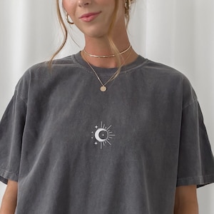 Może przedstawiać: Oversizowa koszulka w kolorze antracytowym z białym nadrukiem w kształcie półksiężyca i gwiazdy. Koszulka ma luźny krój i lekko opadające ramiona.