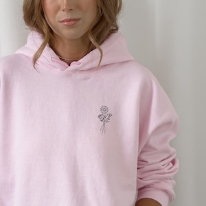 Puede incluir: Una sudadera con capucha rosa con un pequeño diseño floral bordado en la parte delantera. La sudadera tiene una capucha con cordón y un bolsillo canguro.