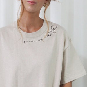 May include: An oversized beige t-shirt with a simple floral design and the text "you are beautiful" on the front.