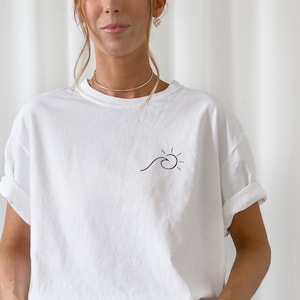 Op de afbeelding: Wit T-shirt met een eenvoudige zwarte lijntekening van een golf en zon op de linker borst.