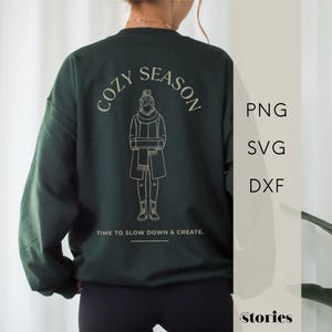 May include: Dark green sweatshirt with the words "COZY SEASON" arched above a line drawing of a person wearing a scarf and coat. Below the drawing is the text "TIME TO SLOW DOWN & CREATE."