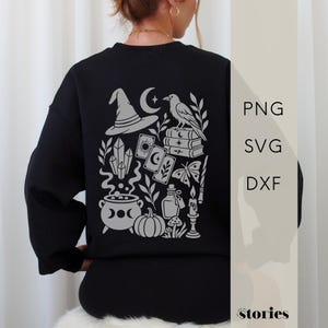 Könnte beinhalten: Ein schwarzes Langarm-Sweatshirt mit einer großen hellgrauen Grafik auf der Rückseite. Das Design zeigt einen Hexenhut, eine Mondsichel, einen Raben, Bücherstapel, Kristalle, Tarotkarten, einen Schmetterling, einen Kessel, einen Kürbis, eine Flasche und eine Kerze. Text: PNG, SVG, DXF.