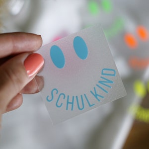 Könnte beinhalten: Ein transparenter Aufkleber mit einem blauen Smiley und dem Text "SCHULKIND" in blau.
