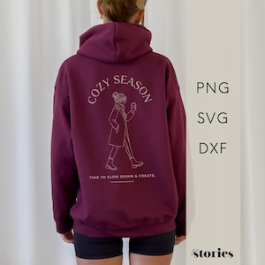 Puede incluir: Sudadera con capucha burdeos con un gráfico blanco en la espalda. El gráfico incluye las palabras "COZY SEASON" sobre un dibujo lineal de una persona sosteniendo una taza. Debajo del gráfico está el texto "TIME TO SLOW DOWN & CREATE".