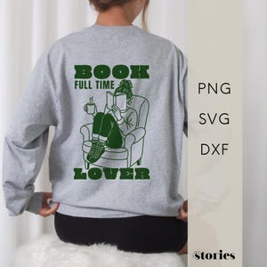 Puede incluir: Sudadera gris con las palabras "BOOK FULL TIME LOVER" en verde. Una ilustración verde muestra a una persona leyendo un libro en un sillón, sosteniendo una taza. La sudadera tiene mangas largas.