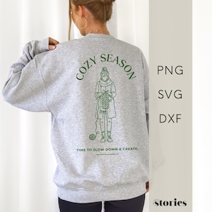 Könnte beinhalten: Hellgraues Sweatshirt mit grünem Aufdruck auf dem Rücken. Der Aufdruck zeigt die Worte "COZY SEASON" und eine Illustration einer strickenden Person. Darunter steht "TIME TO SLOW DOWN & CREATE."