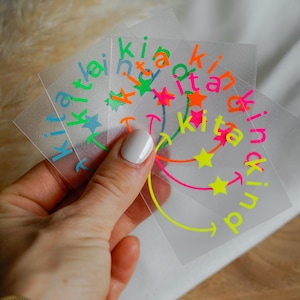 Könnte beinhalten: Eine Hand hält mehrere transparente quadratische Aufkleber. Jeder Aufkleber zeigt das Wort "kind" in einem kreisförmigen Design mit neongrünen, orangefarbenen, rosa und blauen Farben. Das Design beinhaltet Sterne und Smileys.