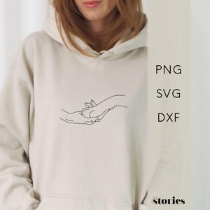 Könnte beinhalten: Ein beigefarbener Kapuzenpullover mit einer schwarzen Strichzeichnung einer menschlichen Hand, die eine Hundepfote hält. Der Text PNG, SVG und DXF ist vertikal auf der rechten Seite gedruckt, und das Wort stories steht unten rechts.
