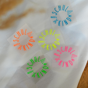 Op de afbeelding: Vier strijkbare vinyl patches met de tekst "School is Kind" in een cirkel met schoolbenodigdheden rond de tekst. De patches zijn in verschillende kleuren: blauw, groen, oranje en roze.