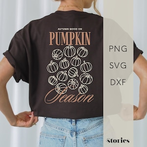 Könnte beinhalten: Ein dunkelbraunes T-Shirt mit einem Aufdruck auf der Rückseite, der den Text "AUTUMN MOOD ON PUMPKIN SEASON" in hellem Pfirsichton zeigt. Das Design enthält eine Ansammlung von fünfzehn weißen Kürbis-Strichzeichnungen in der Mitte.