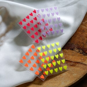 May include: Three sheets of clear adhesive paper with heart-shaped stickers. The stickers are in three colours: red, yellow, and purple.