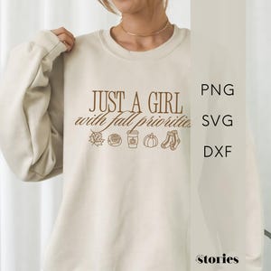 May include: A cream-colored crewneck sweatshirt featuring brown text that reads "JUST A GIRL with fall priorities" above line art icons of a maple leaf, a cinnamon roll, a coffee cup, a pumpkin, and a pair of socks. The design is centered on the chest.