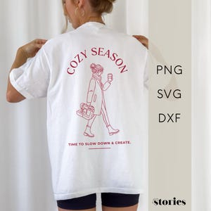 以下が含まれることがあります： 白いTシャツに赤いグラフィック。グラフィックには「COZY SEASON」の文字と、コーヒーカップとバッグを持った人のイラストが描かれています。「TIME TO SLOW DOWN & CREATE」の文字が下にあります。
