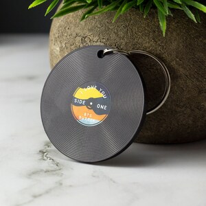 Könnte beinhalten: Schwarzer Vinyl-Schlüsselanhänger mit einem bunten Design, auf dem "I Love You" und "Side One" steht, sowie eine kleine Grafik eines Plattenspielers.