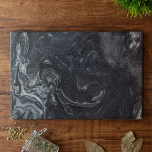 Puede incluir: Una tabla de cortar de m&aacute;rmol negro con un patr&oacute;n blanco en espiral. La tabla es rectangular y tiene una superficie lisa.
