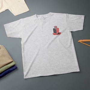 K&ouml;nnte beinhalten: Hellgraues T-Shirt mit einem kleinen Brustaufdruck in Rot, Schwarz und Grau mit dem Wort "TRIUMPH" in Rot. Weitere gefaltete T-Shirts und ein Holzkleiderb&uuml;gel sind ebenfalls zu sehen.