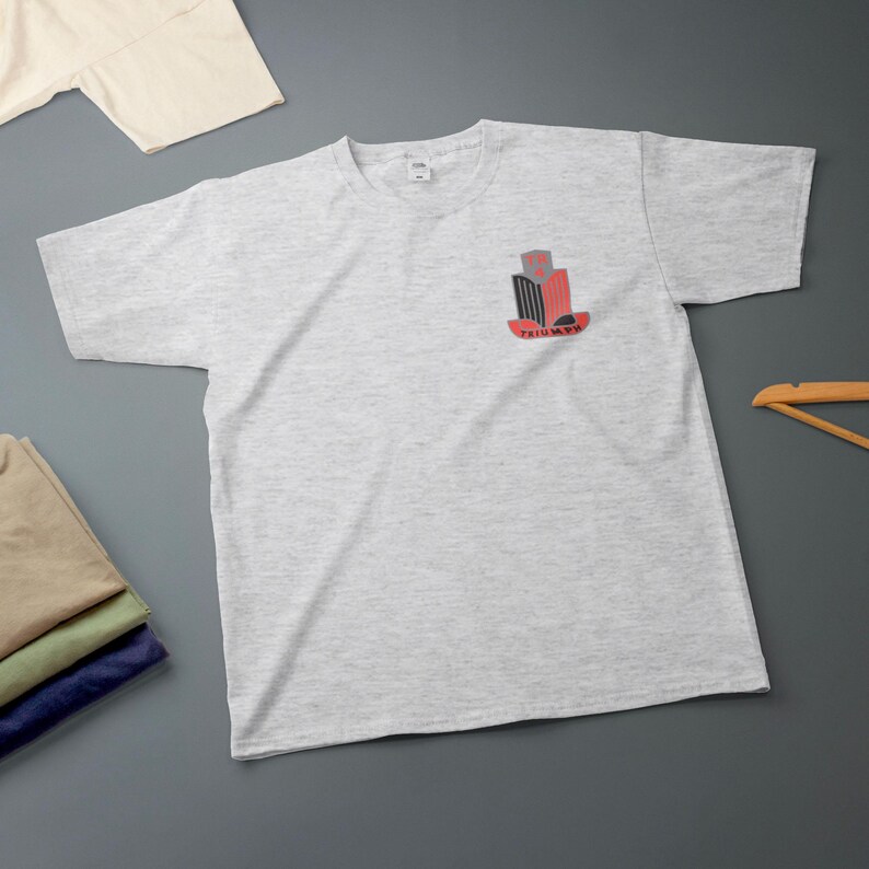 K&ouml;nnte beinhalten: Hellgraues T-Shirt mit einem kleinen Brustaufdruck in Rot, Schwarz und Grau mit dem Wort "TRIUMPH" in Rot. Weitere gefaltete T-Shirts und ein Holzkleiderb&uuml;gel sind ebenfalls zu sehen.