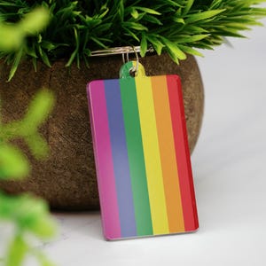 Könnte beinhalten: Ein rechteckiger Schlüsselanhänger mit Regenbogenmuster. Er hat vertikale Streifen in Rot, Orange, Gelb, Grün, Blau und Lila. Er ist an einem silbernen Metallring befestigt.