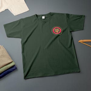 Könnte beinhalten: Dunkelgrünes T-Shirt mit einem kreisförmigen Emblem auf der linken Brust. Das Emblem zeigt Text und ein Wappen-Design in Rot, Gold und Grün. Zusätzliche gefaltete Hemden und ein Kleiderbügel befinden sich im Hintergrund.