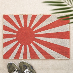 May include: A rectangular doormat featuring a red sunburst design with white rays. The mat is made of a textured material and is placed on a light-colored surface. Camouflage sneakers are visible at the bottom of the image.