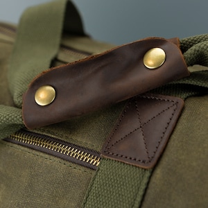May include: Close-up of a green canvas duffel bag with brown leather accents. The bag has a zipper closure and a leather handle with brass accents.