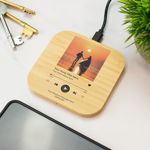 May include: A square, bamboo wireless charger featuring a sunset image and a music player interface. The image shows a couple holding hands on a beach. Text includes "Your Song Title Here" and "Your Artist Name Here."