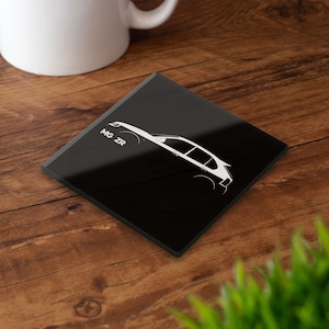 Puede incluir: Posavasos cuadrado negro con un contorno blanco de un coche y el texto "MG ZR". El posavasos está sobre una superficie de madera, con una taza blanca en el fondo.