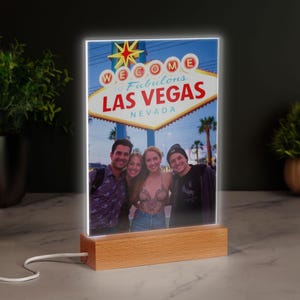 May include: A lit photo display showing four people posing in front of the "Welcome to Fabulous Las Vegas Nevada" sign. The display has a wooden base and a bright, white light around the photo.