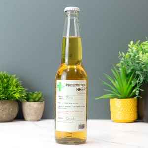 May include: A glass bottle of beer with a white label that reads "Prescription Beer". The label has a green cross and the text "Patient Name: Thomas". The label also includes instructions for taking the beer, such as "Drink one 330ml dose per day." and "In extreme cases, drink entire care prescription."