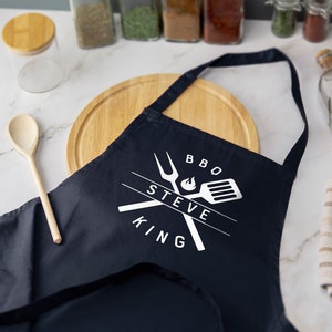 May include: A navy blue apron with white text that reads "BBQ STEVE KING" and a graphic of crossed grilling tools with a flame.