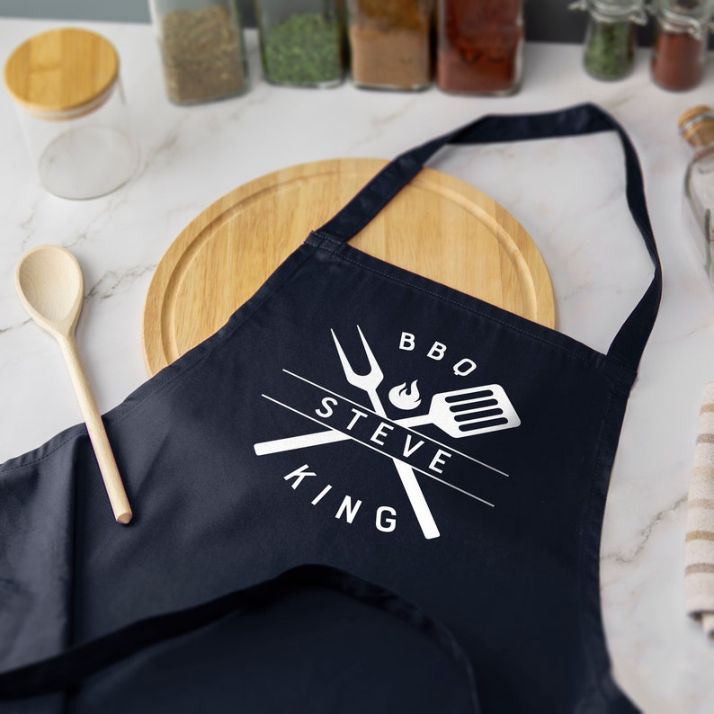 May include: A navy blue apron with white text that reads "BBQ STEVE KING" and a graphic of crossed grilling tools with a flame.